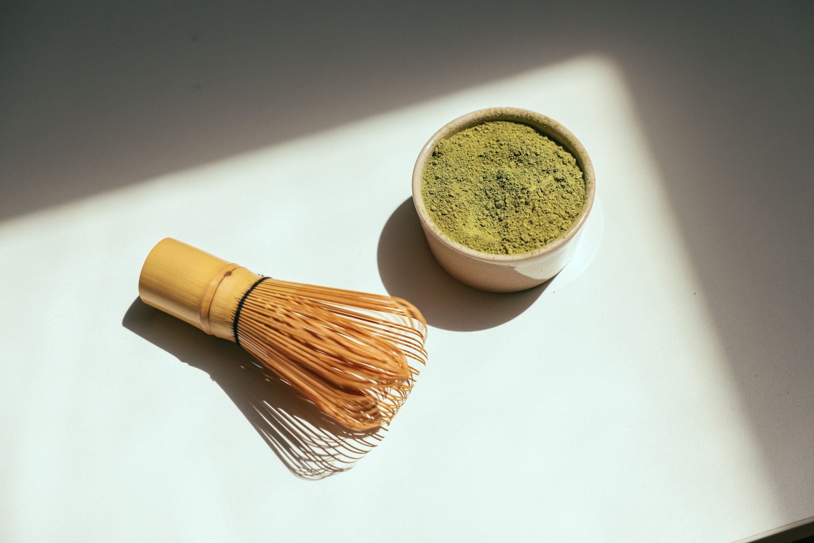 A bamboo matcha whisk beside a bowl of powdered matcha, illuminated by natural sunlight.